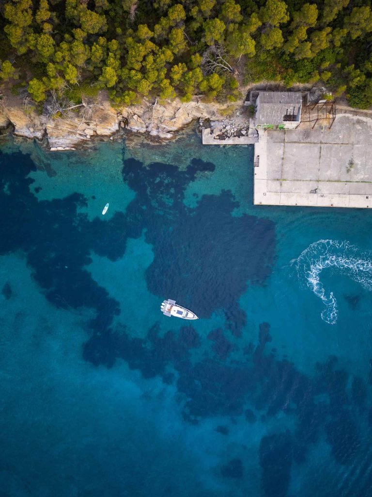 A stunning aerial shot of Paralos yacht anchored near a rugged cliffside with clear, turquoise waters below and lush green trees above