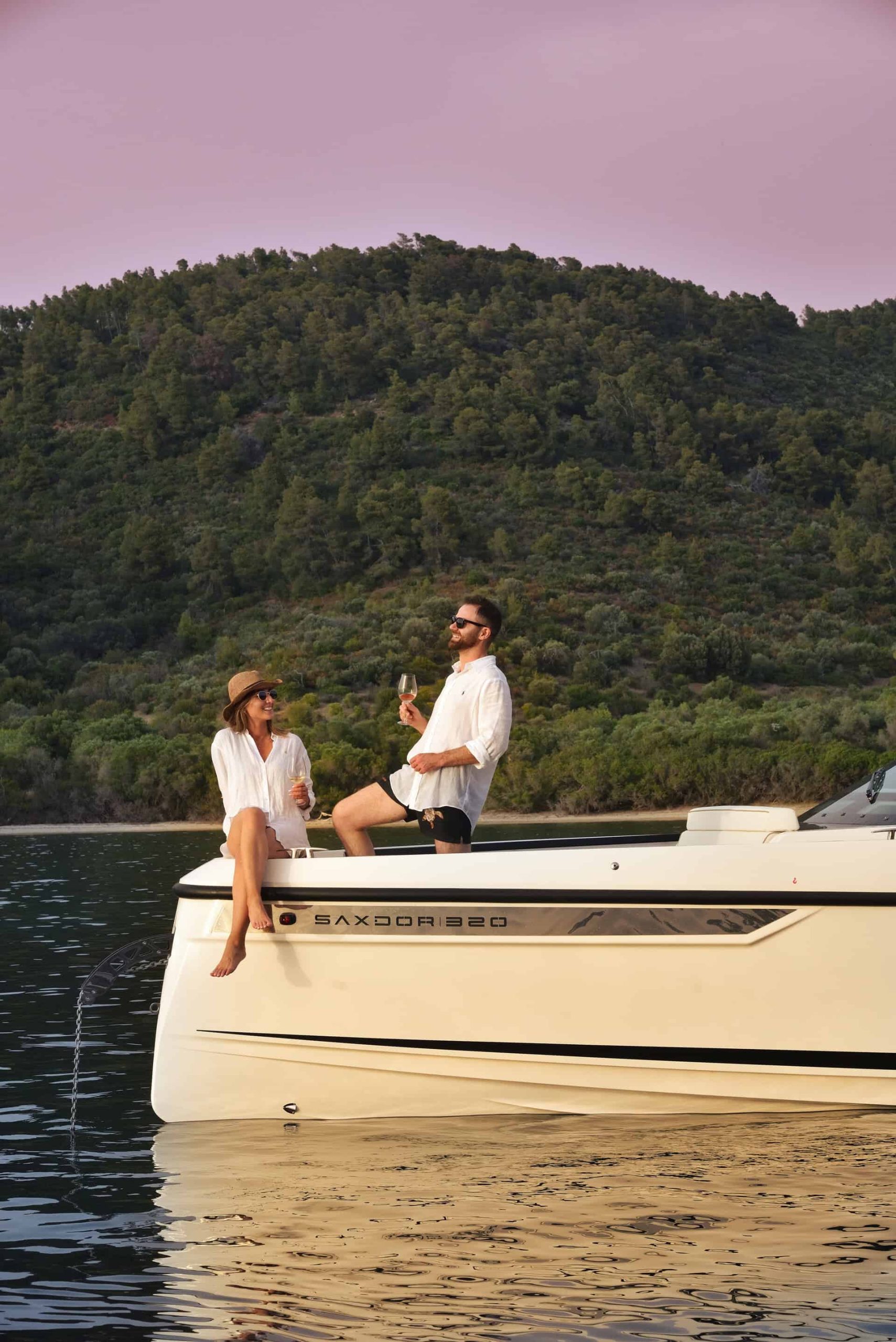 A couple enjoying drinks on the bow of Paralos yacht at sunset, with lush green hills in the background