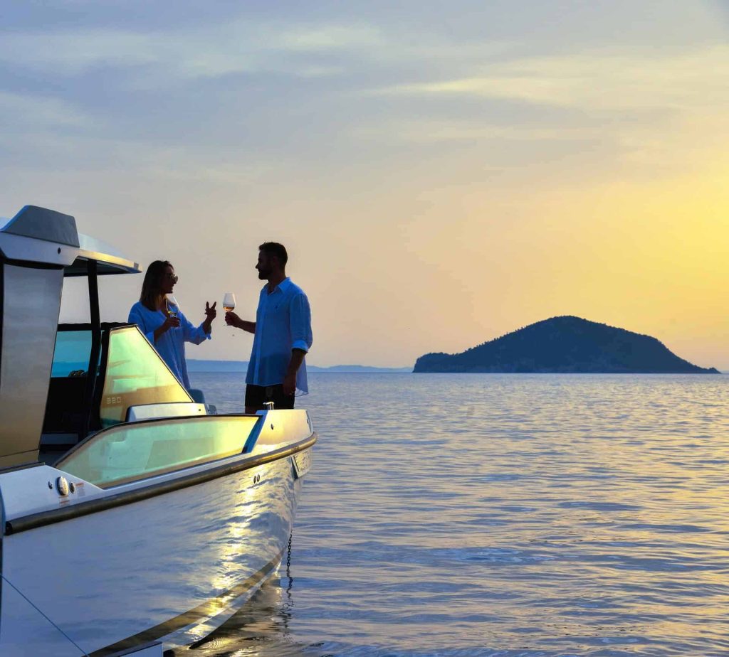 A couple toasting with drinks on the deck of the Paralos boat at sunset, with a scenic island in the background