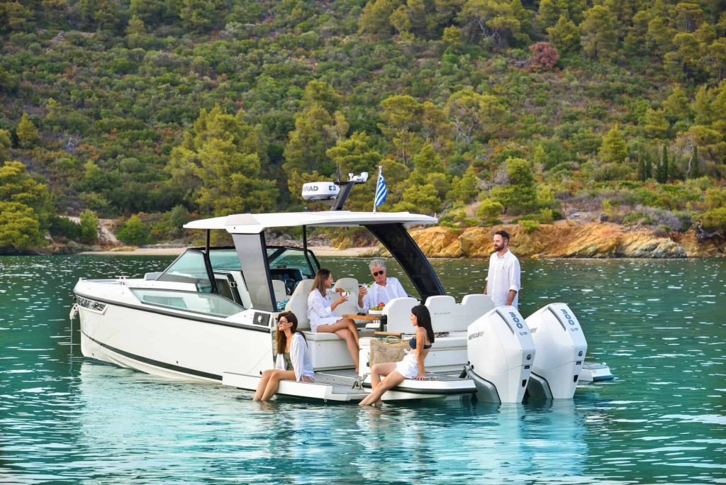 A group of friends and family relax on the deck of the Saxdor 320 GTO 'Paralos' yacht, anchored in a picturesque cove surrounded by lush greenery in Halkidiki