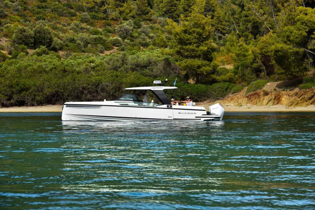 A luxury yacht, Paralos, anchored close to a scenic shoreline with lush greenery in the background, with people enjoying time on board