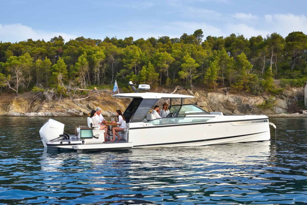 A luxury motorboat anchored near a rocky, forested shoreline with a group of people enjoying a meal on the deck