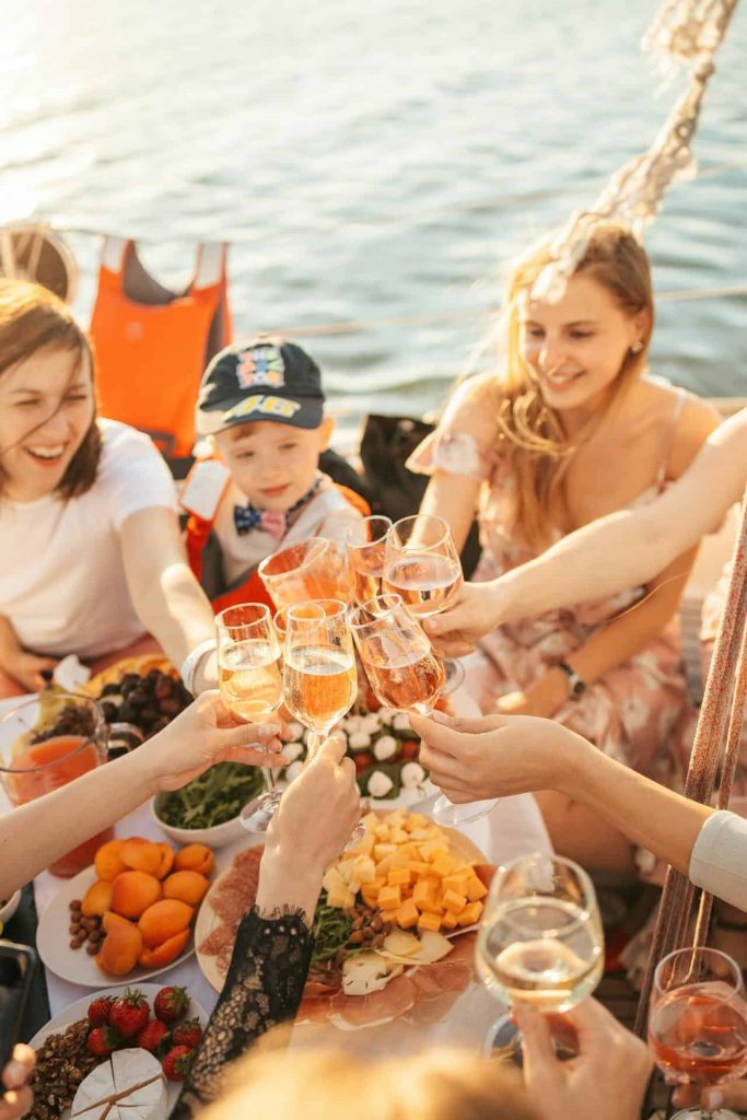 A family and friends enjoying a toast with champagne glasses on a yacht, surrounded by a variety of delicious foods, including fruits, cheese, and meats, with a beautiful water backdrop