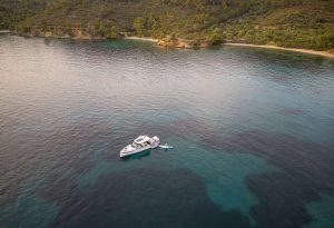 Aerial view of Paralos yacht anchored in a calm bay with a paddleboarder next to it, surrounded by serene hills and clear water
